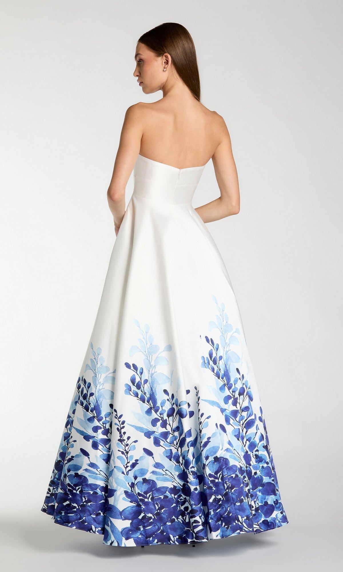 A woman stands with her back to the camera against a plain background, wearing the Mac Duggal 56316 long formal dress—a strapless Mikado ballgown featuring a blue floral pattern along the lower half.